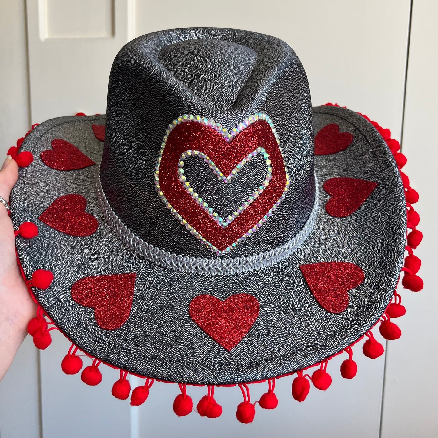 Gray hat with red heart designs and pom-poms held against a white wall.