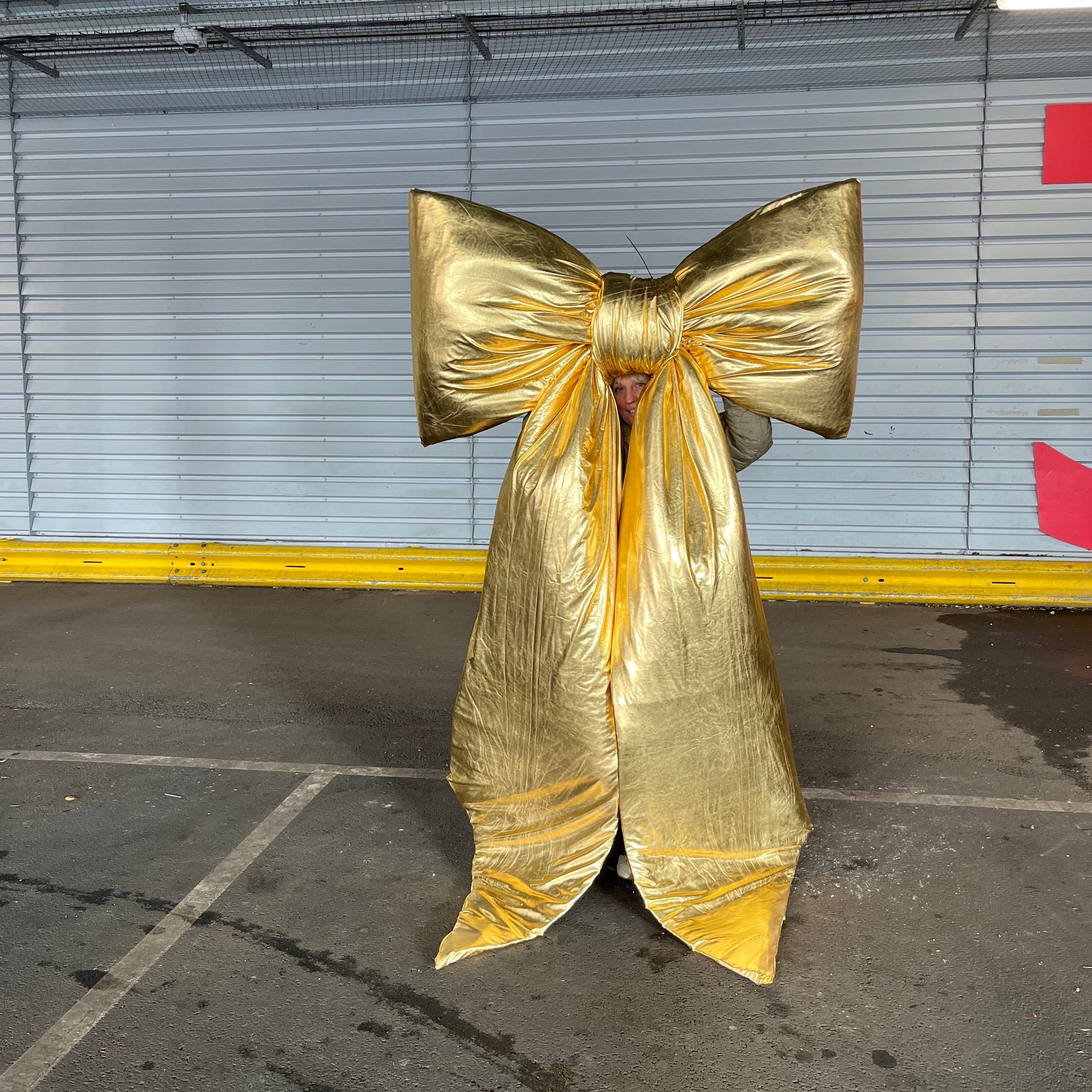 A person holding up a shiny metallic gold large bow, standing in an indoor parking garage with a gray metal roll-up door behind them.