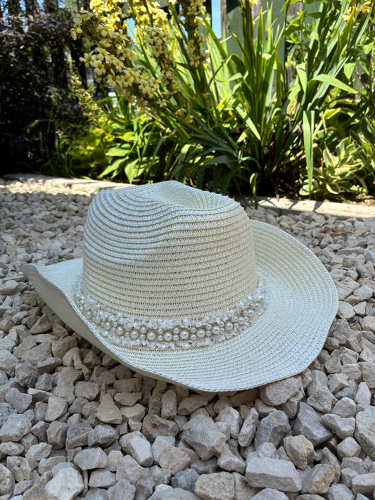 White straw hat with decorative trim on a bed of small stones with plants in the background