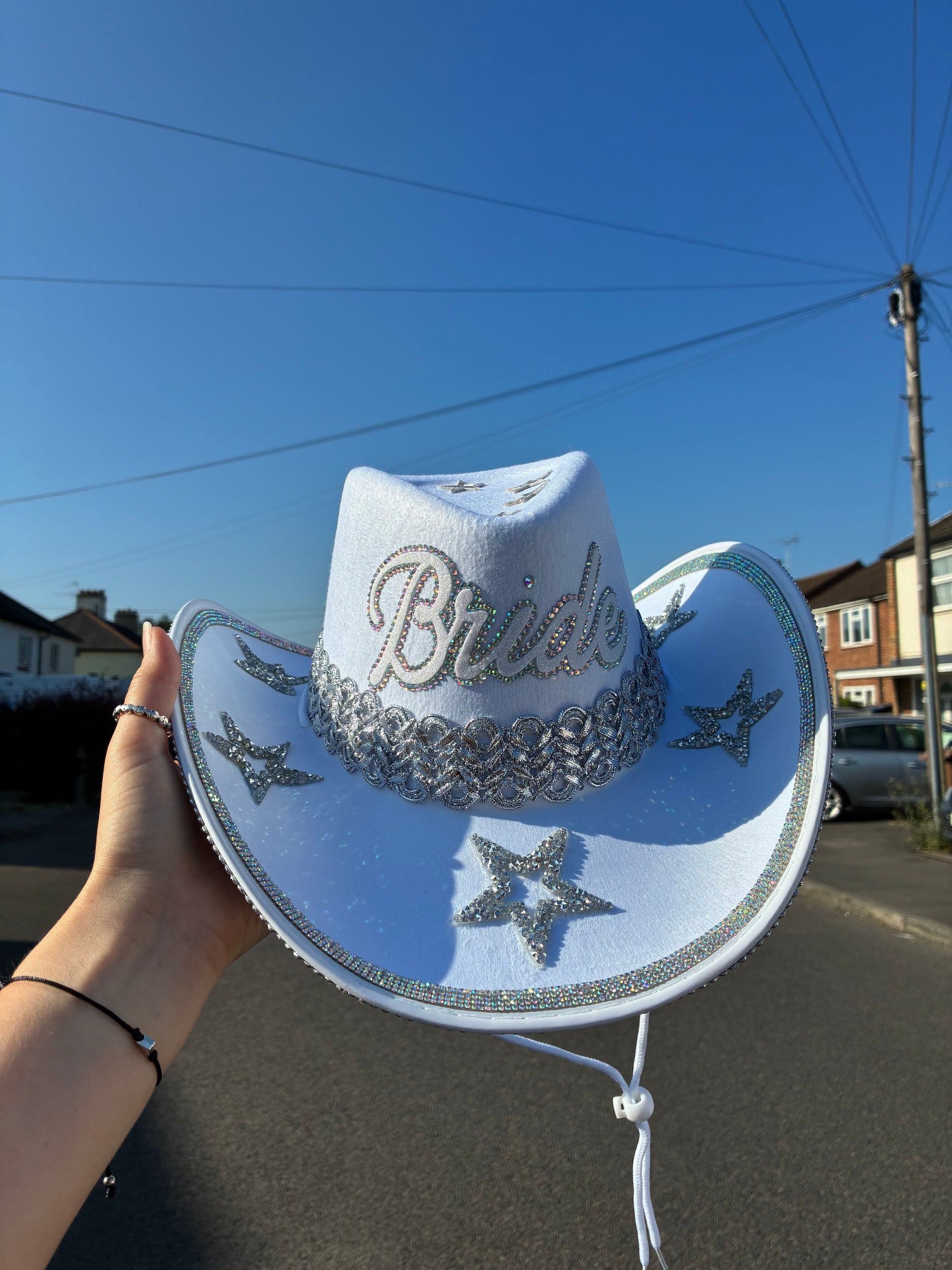 A white cowboy hat with silver rhinestone embroidery spelling 'Bride' and star motifs, held up by a hand against an outdoor street background with a blue sky.