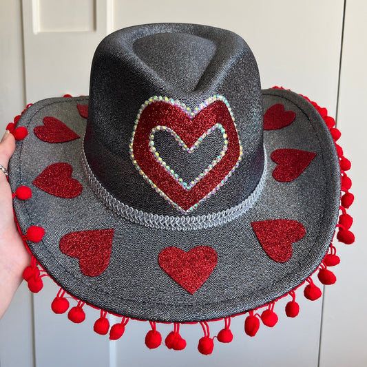 Gray hat with red heart designs and pom-poms held against a white wall.